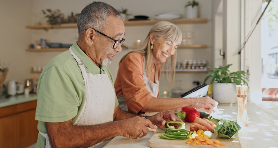 A imagem mostra um casal de idosos cozinhando juntos em uma cozinha bem iluminada e acolhedora. Ambos parecem felizes e relaxados, sorrindo enquanto preparam alimentos frescos.