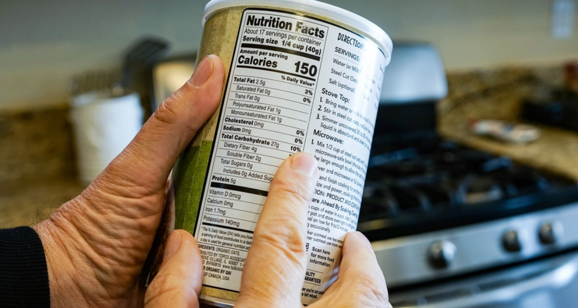 A imagem mostra uma pessoa segurando e lendo o rótulo nutricional de um produto alimentar, provavelmente um tipo de aveia ou cereal. A embalagem está em destaque, com foco na tabela de informações nutricionais.
