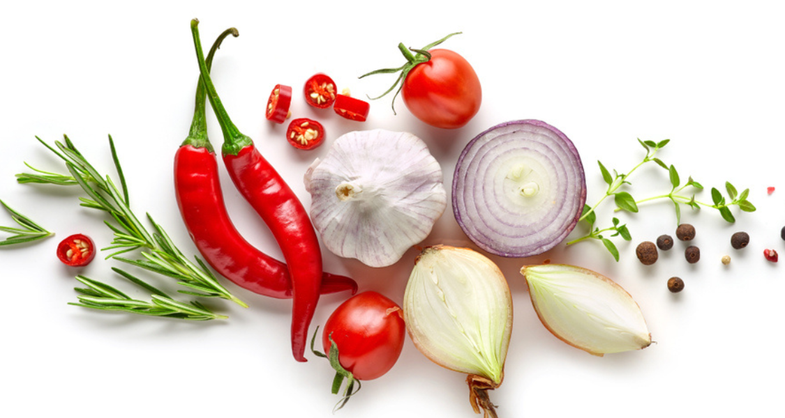 A imagem mostra uma bela e colorida seleção de temperos naturais e vegetais frescos dispostos sobre uma superfície branca. Estes ingredientes são ótimas alternativas para reduzir o uso de sal (sódio) nos alimentos, pois realçam o sabor de forma natural e saudável.