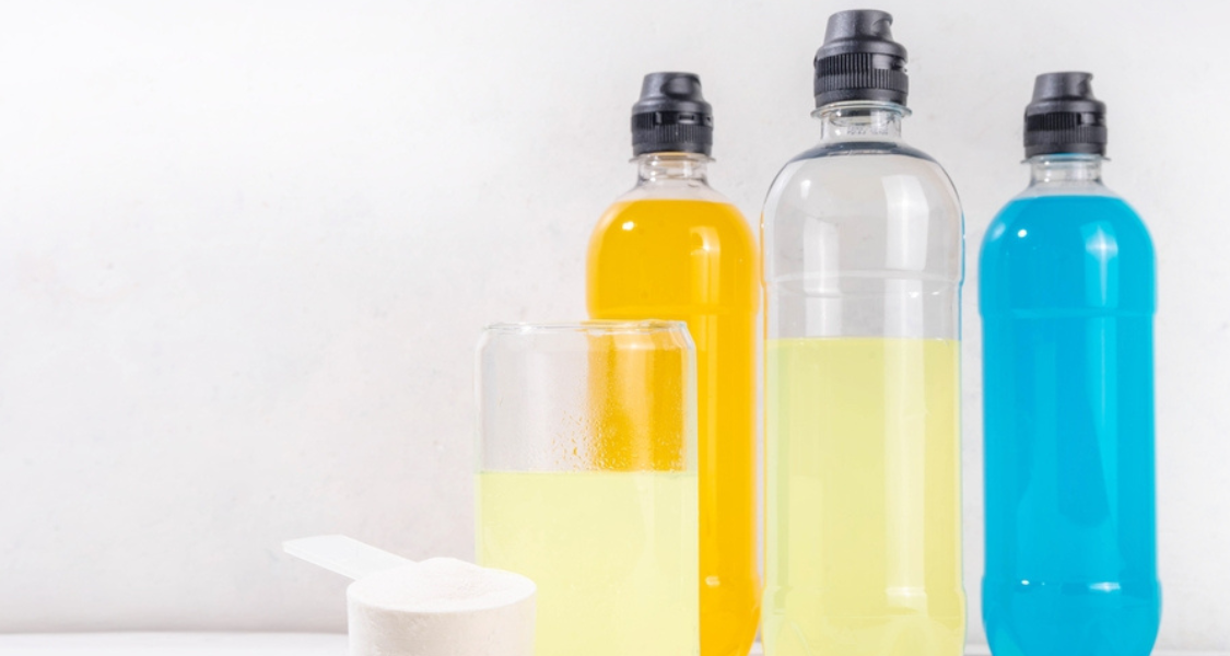 A imagem mostra três garrafas esportivas contendo líquidos coloridos e uma bebida em um copo, ao lado de um medidor com pó branco, provavelmente um suplemento ou bebida isotônica em pó antes da diluição.