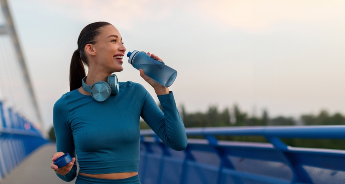 A imagem mostra uma mulher jovem em um ambiente ao ar livre, provavelmente em uma ponte ou passarela urbana, bebendo água de uma garrafa esportiva. A cena transmite energia, bem-estar e estilo de vida saudável.