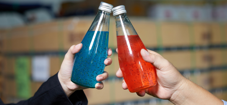 A imagem mostra duas pessoas brindando com garrafas de vidro contendo bebidas coloridas com sementes em suspensão. As bebidas parecem ser águas funcionais ou refrescos com sementes de chia ou manjericão, bastante populares por seus benefícios à saúde.