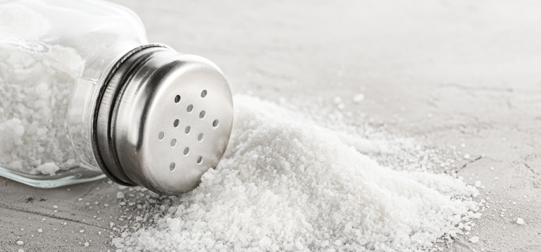 A imagem mostra um saleiro de vidro e tampa metálica deitado, com sal grosso branco espalhado sobre uma superfície clara (possivelmente uma bancada ou mesa de pedra).