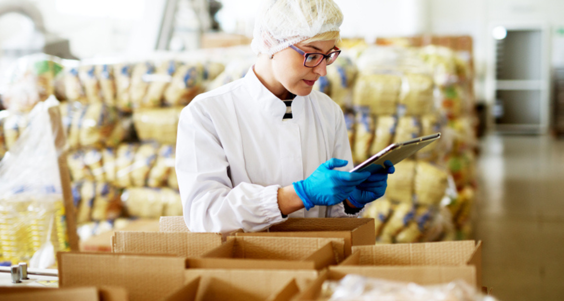 A imagem mostra uma profissional da indústria alimentícia trabalhando em um ambiente de armazenagem seca ou linha de embalagem de alimentos não perecíveis.