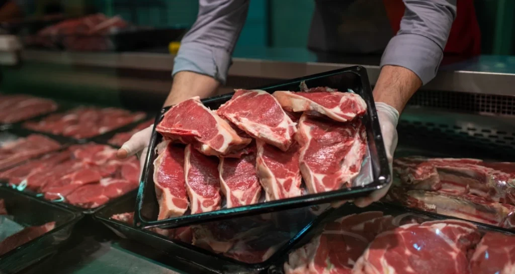 A imagem mostra uma bandeja com cortes de carne bovina crua, exposta em um balcão refrigerado de açougue ou frigorífico. Um funcionário, vestindo luvas descartáveis e camisa social com mangas arregaçadas, segura a bandeja com ambas as mãos, apresentando o produto ao cliente ou organizando a vitrine.