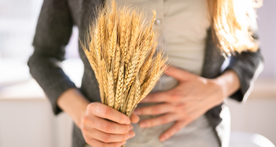 A imagem mostra uma pessoa segurando um feixe de trigo em uma mão enquanto a outra mão está sobre a região abdominal, sugerindo algum desconforto ou dor.