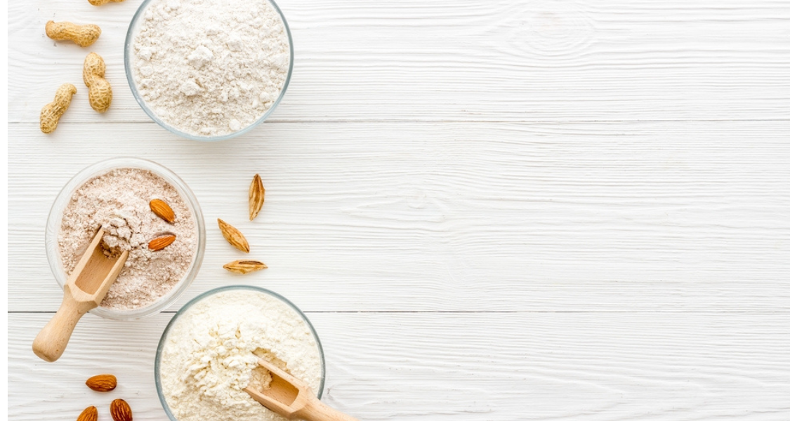 A imagem mostra três tigelas de vidro contendo diferentes tipos de farinhas dispostas sobre uma superfície de madeira branca. Cada tigela contém um tipo diferente de farinha, aparentemente de oleaginosas ou sementes