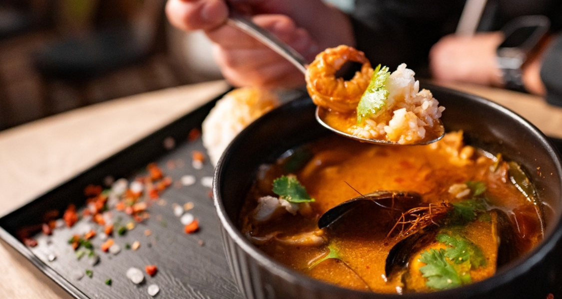 A imagem mostra uma pessoa a desfrutar de um prato de sopa ou guisado de marisco, com foco numa colher cheia de comida a ser levantada do prato.