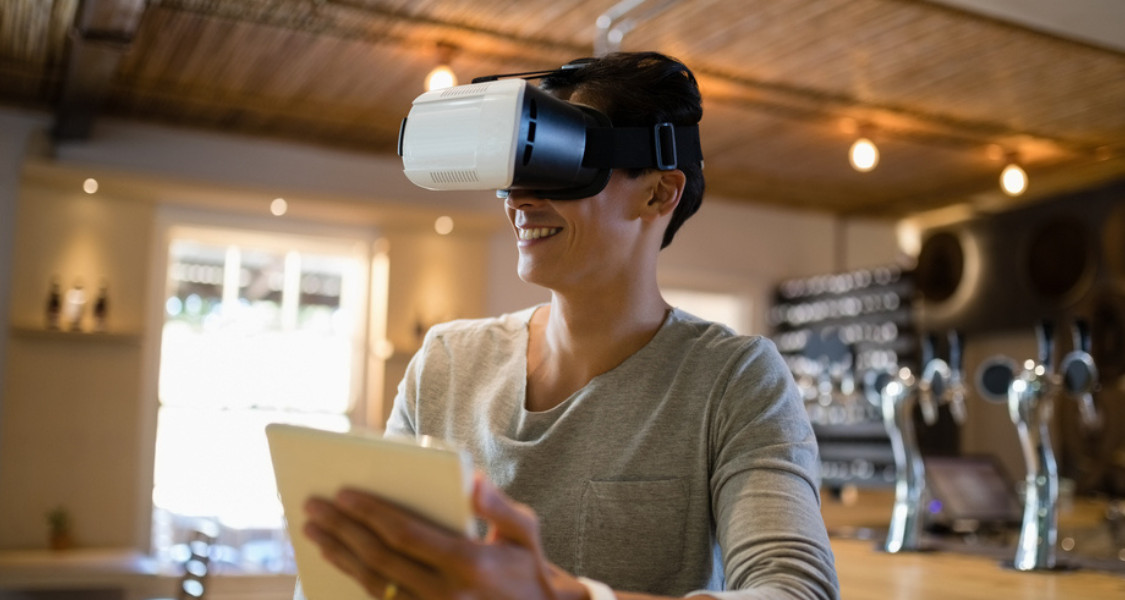 A imagem mostra uma pessoa sorrindo e usando óculos de realidade virtual (VR) brancos e pretos, imersa numa experiência digital. A pessoa, que tem cabelo escuro e curto, veste uma t-shirt cinzenta simples e segura um tablet branco na mão esquerda, que está ligeiramente desfocado em primeiro plano.
