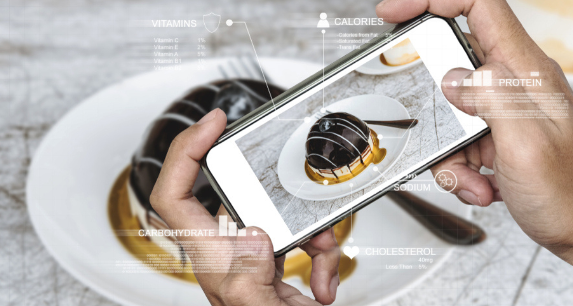 A imagem mostra uma pessoa a segurar um smartphone na horizontal, a tirar uma fotografia de uma sobremesa num prato. O ecrã do telemóvel exibe a imagem nítida da mesma sobremesa que está na mesa, sugerindo que a fotografia está a ser capturada em tempo real.