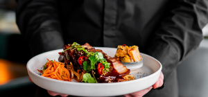 A imagem mostra um close-up de um prato de comida gourmet lindamente apresentado, sendo segurado por uma pessoa vestindo um uniforme escuro (possivelmente um garçom ou chef), cujo tronco é apenas visível no fundo desfocado.