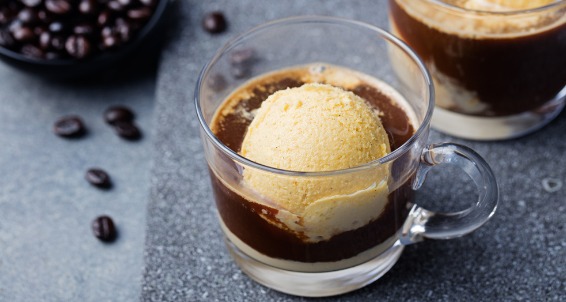 A imagem é uma fotografia de close-up, com foco raso, mostrando uma sobremesa de café clássica, provavelmente um Affogato, em um cenário que remete a uma cafeteria.