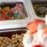 A imagem é um close-up que foca em uma variedade de acompanhamentos e coberturas (toppings) para sorvetes ou iogurtes congelados, tipicamente encontrados em uma barra de self-service.