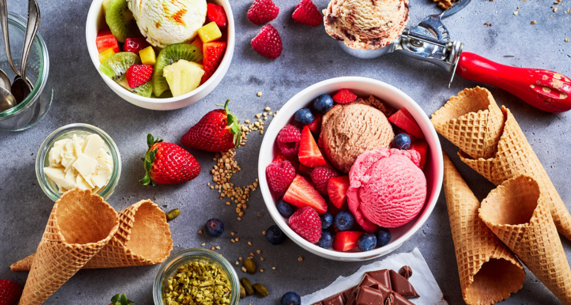 A imagem é uma fotografia de ângulo superior (plano flat lay) que apresenta uma variedade rica e colorida de sorvetes, frutas, coberturas e acompanhamentos, dispostos sobre uma superfície escura e texturizada (que se assemelha a concreto ou ardósia). A cena sugere um banquete de verão ou um menu gourmet de sobremesas.