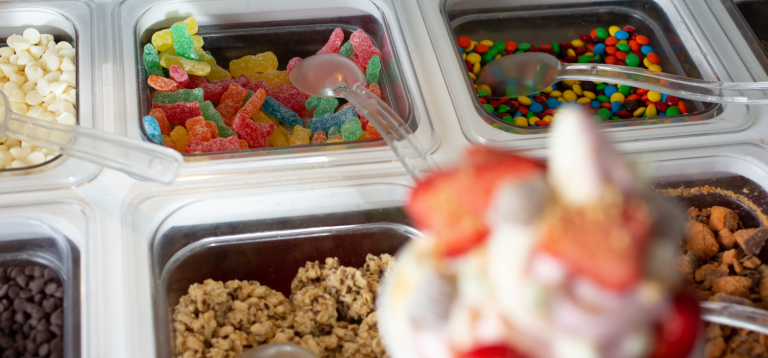 A imagem é um close-up que foca em uma variedade de acompanhamentos e coberturas (toppings) para sorvetes ou iogurtes congelados, tipicamente encontrados em uma barra de self-service.