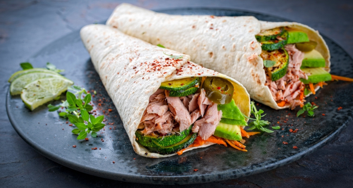 A imagem mostra dois wraps dispostos em um prato escuro. Eles estão recheados com pedaços de atum, abacate, rodelas de abobrinha grelhada, cenoura ralada e pimenta jalapeño.