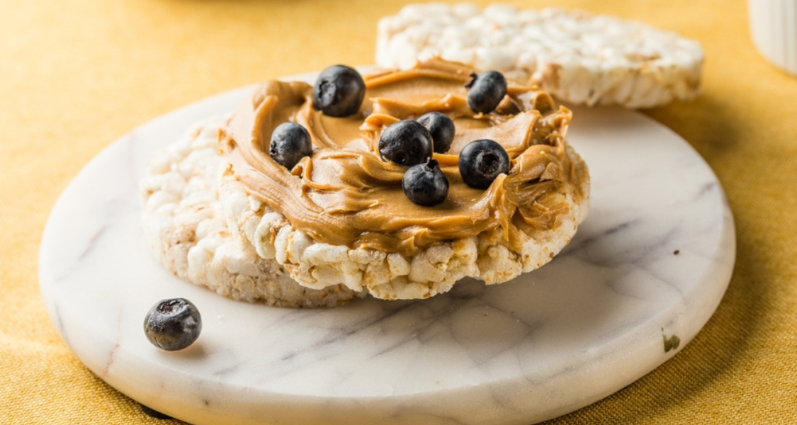 Na imagem, há um lanche composto por um biscoito de arroz coberto com pasta de amendoim cremosa e mirtilos frescos no topo.