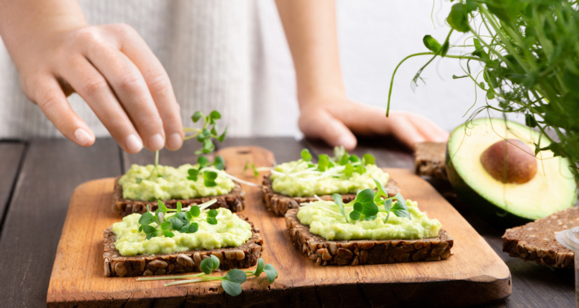 A imagem mostra quatro torradas de pão integral com sementes, dispostas sobre uma tábua de madeira. Cada torrada está coberta com uma pasta cremosa de abacate e finalizada com pequenas folhas verdes frescas, que uma pessoa está cuidadosamente colocando com a mão.