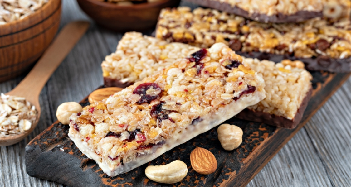 A imagem mostra várias barrinhas de cereal dispostas sobre uma tábua de madeira escura. Elas são feitas de aveia, frutas secas como cranberries e cobertas com uma camada branca ou de chocolate na base. Ao redor, há castanhas e amêndoas espalhadas, além de uma colher de madeira com aveia, reforçando o aspecto natural e nutritivo do lanche.