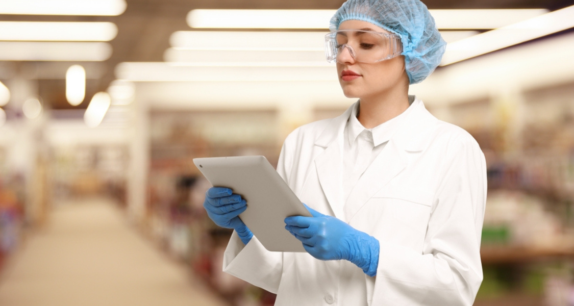 A imagem retrata uma profissional da área de ciência, tecnologia ou controle de qualidade em um ambiente comercial, usando um dispositivo digital para análise ou registro de dados.