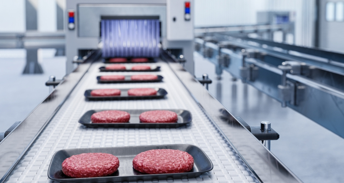 A imagem mostra uma linha de produção automatizada na indústria alimentícia, possivelmente em uma fábrica de processamento de carne. Sobre uma esteira transportadora metálica, há bandejas pretas contendo hambúrgueres de carne crua, dispostos de forma organizada.