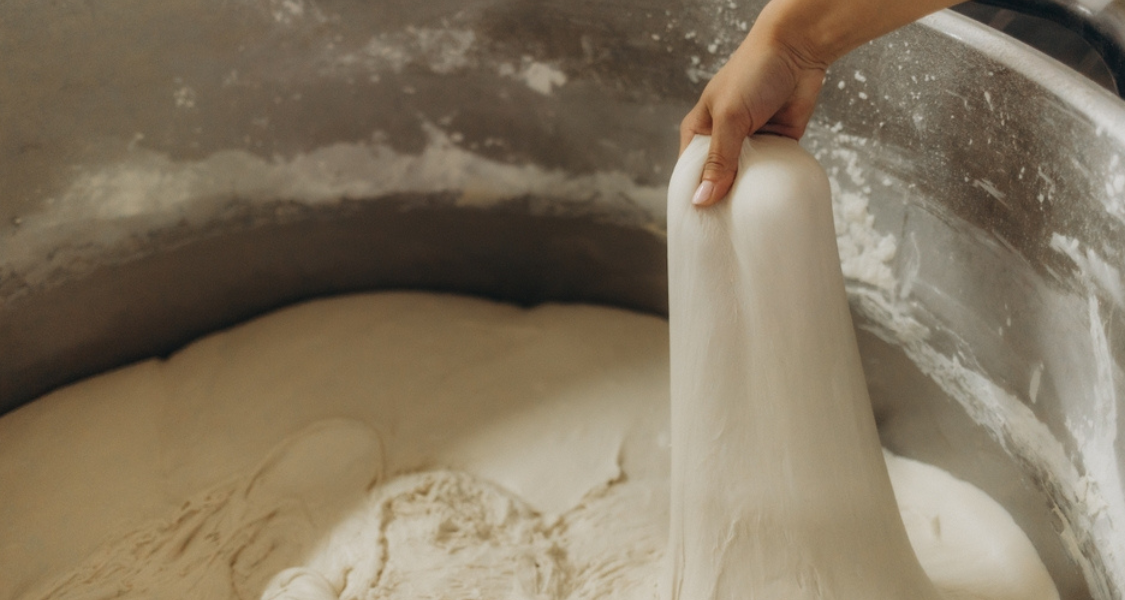 A imagem mostra uma mão puxando uma porção de massa elástica e homogênea de dentro de um grande recipiente metálico.