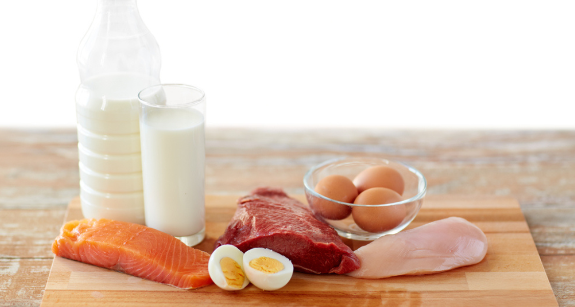 A imagem mostra uma seleção de alimentos ricos em proteína de alta qualidade dispostos sobre uma tábua de madeira. Há um filé de salmão, um pedaço de carne vermelha, um peito de frango cru, ovos inteiros e cozidos, além de uma garrafa e um copo de leite.