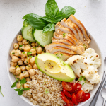 A imagem mostra um prato saudável e colorido com ingredientes frescos e naturais. Nele há peito de frango grelhado fatiado, grão-de-bico cozido, quinoa, abacate, tomates-cereja, fatias de pepino, couve-flor cozida e folhas de manjericão.