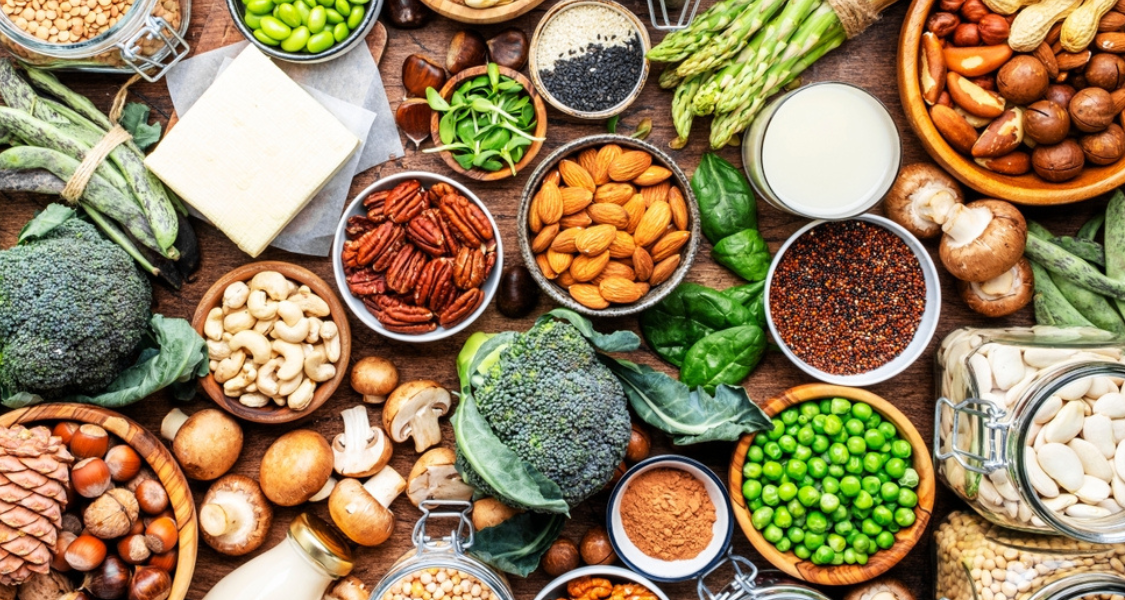 A imagem mostra uma grande variedade de alimentos de origem vegetal ricos em proteína dispostos de forma colorida e organizada sobre uma superfície de madeira. Entre eles há brócolis, ervilhas, aspargos, espinafre, cogumelos, castanhas, amêndoas, nozes, pecãs, cajus, feijões, lentilhas, quinoa, tofu, sementes e leite vegetal.