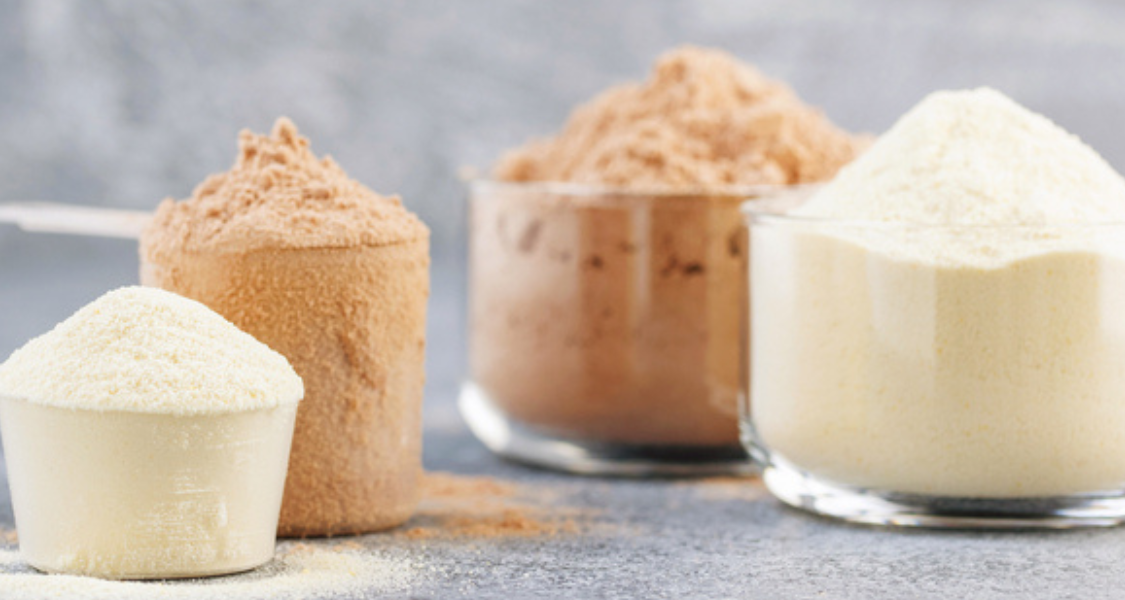 A imagem mostra diferentes tipos de pós proteicos, em tons de bege e marrom, dispostos em copos e medidores transparentes sobre uma superfície cinza.