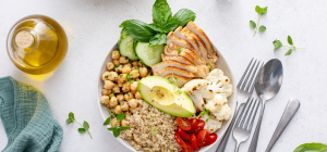 A imagem mostra um prato saudável e colorido com ingredientes frescos e naturais. Nele há peito de frango grelhado fatiado, grão-de-bico cozido, quinoa, abacate, tomates-cereja, fatias de pepino, couve-flor cozida e folhas de manjericão.