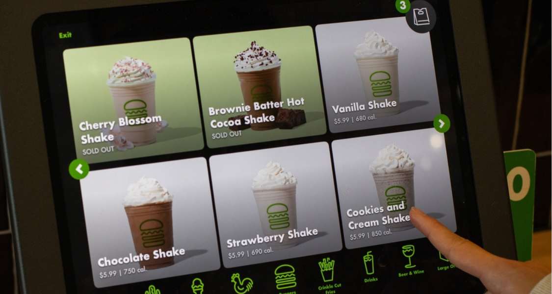 Tela de autoatendimento mostrando cardápio digital de milkshakes com fotos, preços e calorias; uma pessoa seleciona a opção “Cookies and Cream Shake”.
