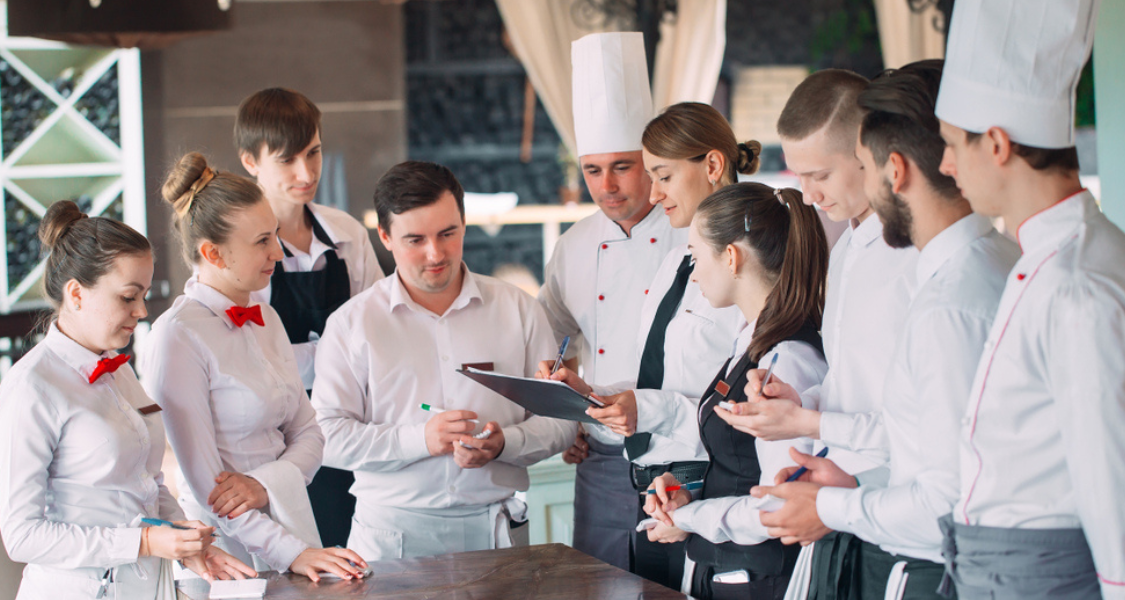 A imagem captura o momento de uma reunião de equipe (briefing ou line-up) em um restaurante ou estabelecimento de food service, focando na comunicação e coordenação entre os membros da equipe.