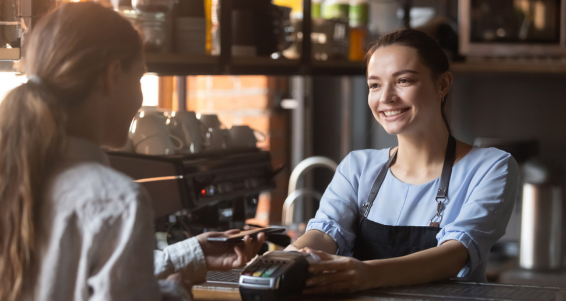 A imagem é um close-up que foca em um momento de transação e interação amigável entre uma atendente e uma cliente no balcão de um café ou bar.