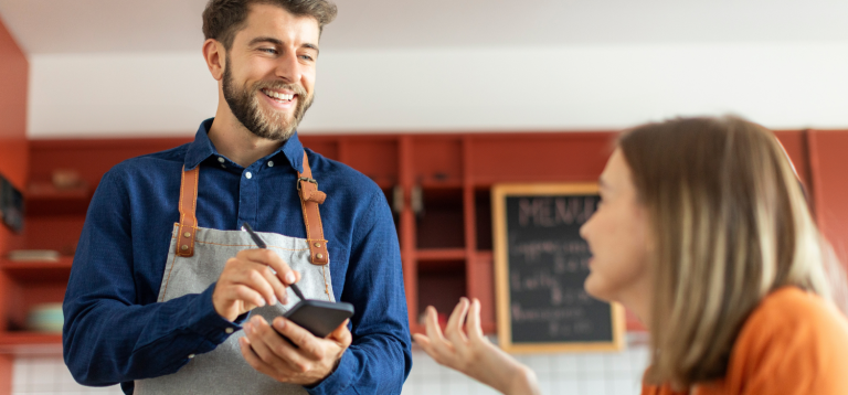 A imagem é uma fotografia que captura um momento de interação positiva entre um funcionário e uma cliente em um ambiente de café ou restaurante moderno e acolhedor.