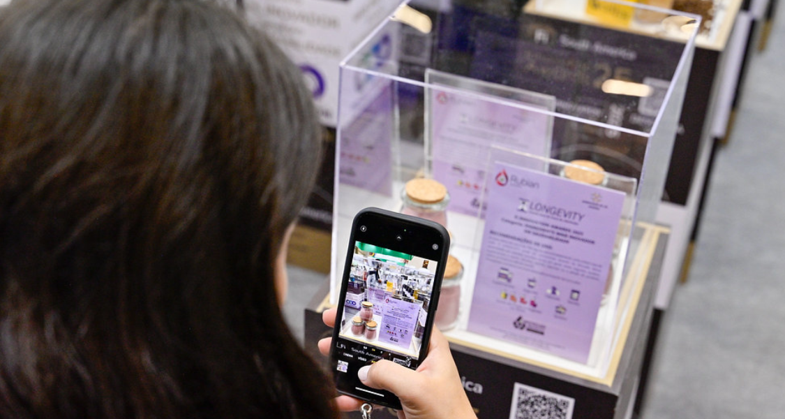 A imagem mostra uma pessoa em um ambiente que parece ser uma feira, exposição ou evento de negócios/ciência, documentando informações com um smartphone.