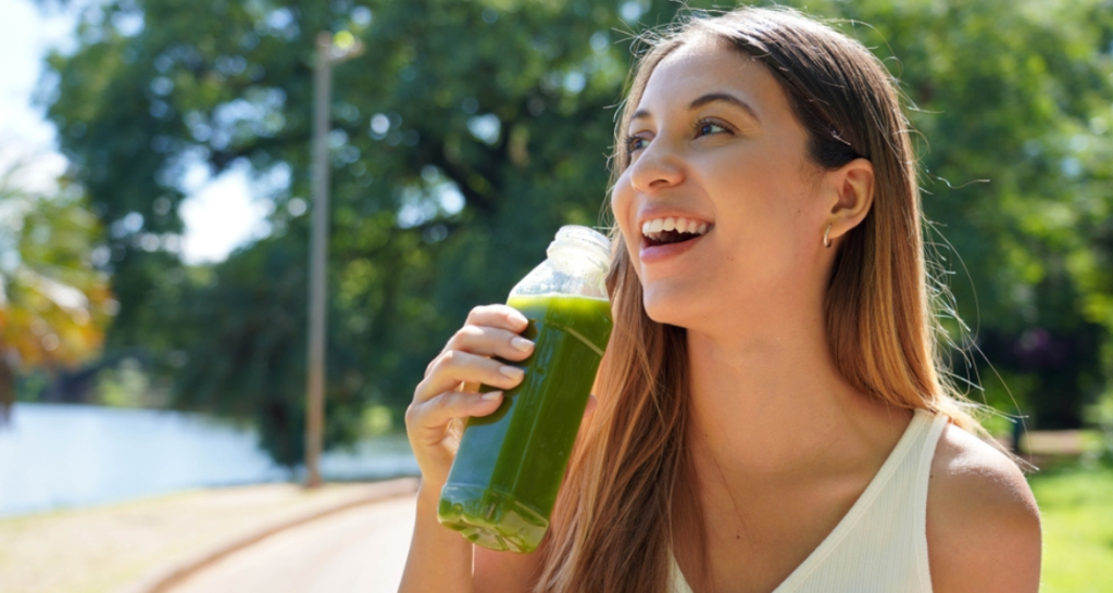 A imagem mostra uma mulher jovem e sorridente desfrutando de uma bebida saudável ao ar livre, em um parque.