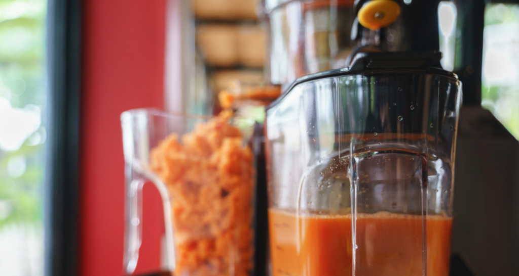 A imagem é um close-up focado em uma máquina de fazer suco, capturando o processo de extração de uma bebida de cor alaranjada, provavelmente suco de cenoura.
