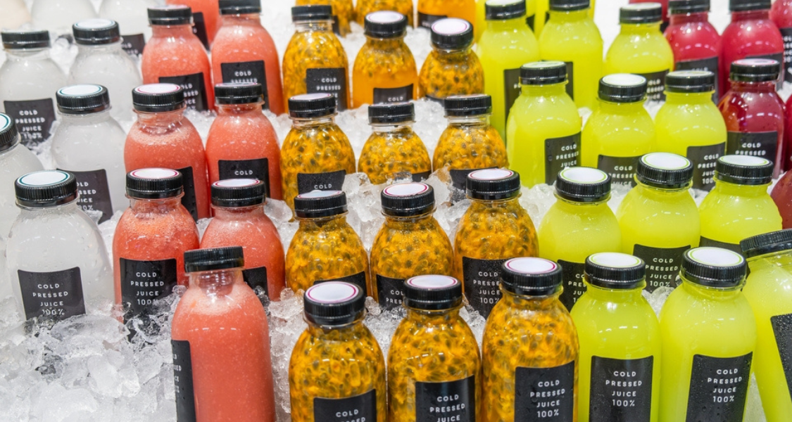 A imagem é um close-up que mostra uma grande variedade de sucos engarrafados expostos e resfriados em gelo, típica de um mercado, feira de alimentos saudáveis ou seção de bebidas frescas.