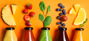 A imagem é uma fotografia de estúdio com uma composição visualmente agradável, apresentando uma linha de garrafas de suco e seus respectivos ingredientes em um fundo laranja vibrante.