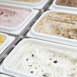 Esta imagem mostra uma visão em close-up de várias bandejas de sorvetes ou gelatos em um freezer de exposição, destacando a variedade de sabores e texturas.