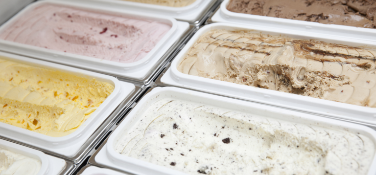 Esta imagem mostra uma visão em close-up de várias bandejas de sorvetes ou gelatos em um freezer de exposição, destacando a variedade de sabores e texturas.