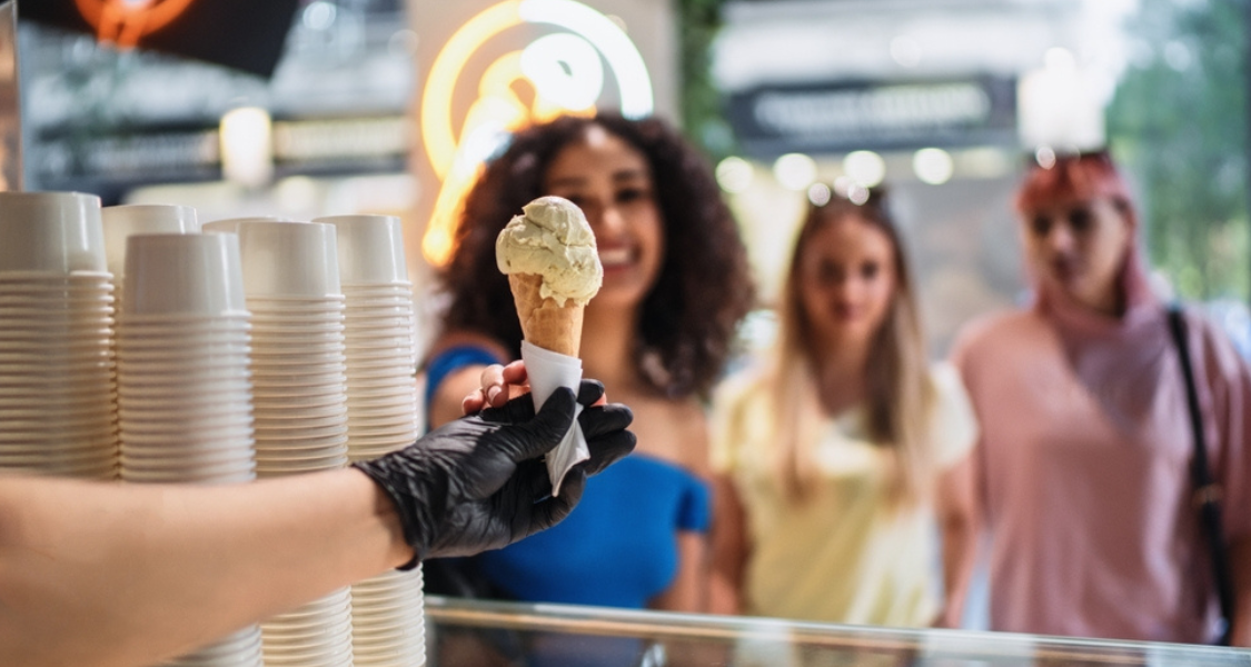 Atendente de sorveteria servindo uma cliente com uma casquinha contendo sorvete.