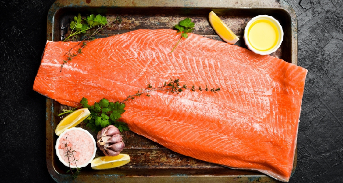 Esta foto, tirada de cima, apresenta um grande filé de salmão cru preparado para o cozimento, disposto artisticamente em uma assadeira rústica.