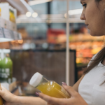 A imagem mostra uma mulher em um corredor de supermercado, aparentemente examinando e comprando bebidas engarrafadas que estão em uma prateleira refrigerada.