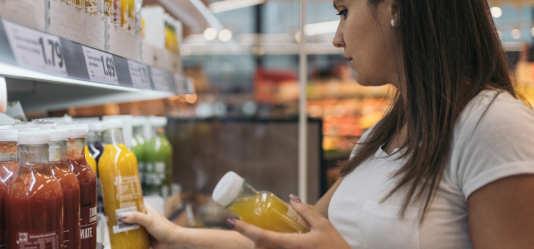 A imagem mostra uma mulher em um corredor de supermercado, aparentemente examinando e comprando bebidas engarrafadas que estão em uma prateleira refrigerada.