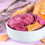 A imagem mostra uma composição gastronômica delicada e bem produzida. Em primeiro plano, há uma tigela pequena de cerâmica azul-clara contendo um sorvete cremoso de cor roxa intensa, com textura suave e aparência artesanal. O sorvete é decorado com pedaços de favo de mel e pequenas flores secas de lavanda, reforçando uma proposta natural e sofisticada.