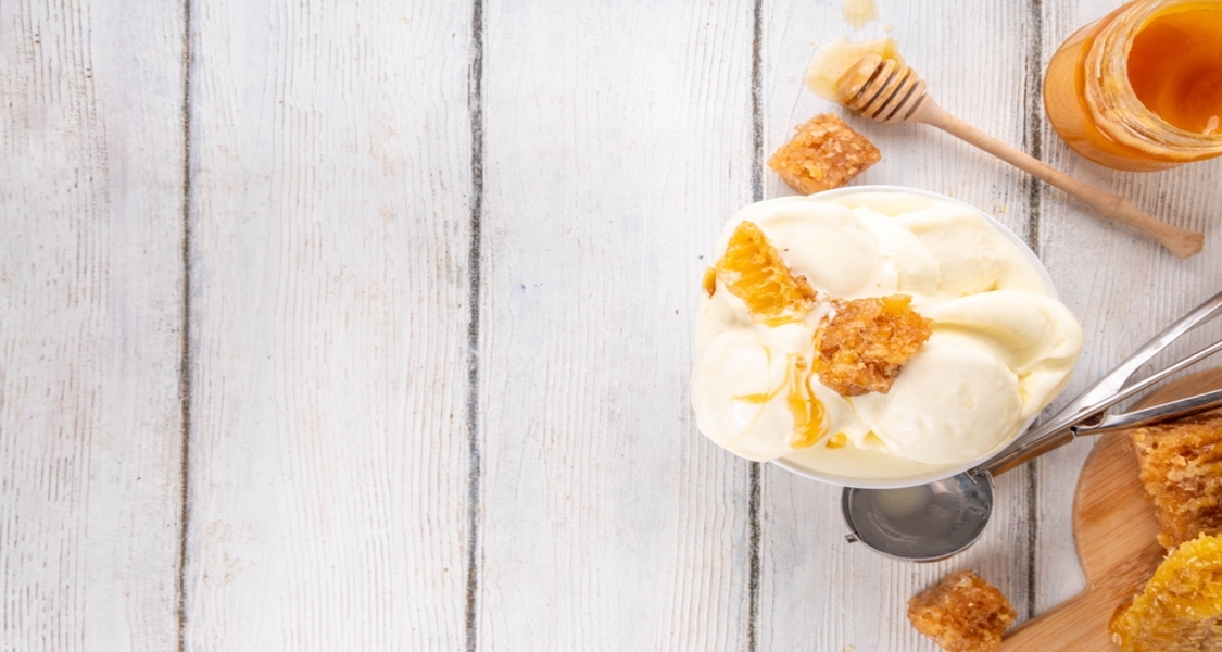 A imagem apresenta uma composição clean e minimalista vista de cima. À direita, há uma tigela com sorvete cremoso de cor clara, com textura lisa e aparência artesanal. Sobre o sorvete, vê-se mel escorrendo e pedaços de favo de mel, que adicionam contraste de cor e textura.