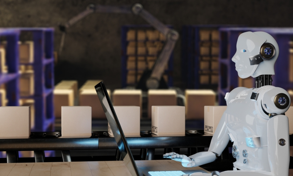 A imagem apresenta um robô humanoide branco sentado diante de um computador em uma linha de produção automatizada. Ao fundo, aparecem caixas sobre uma esteira e um braço robótico, criando um cenário ligado à automação industrial, inteligência artificial e logística automatizada.