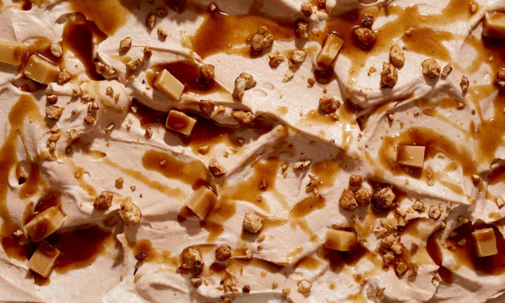 Um close de um sorvete sabor caramelo ou doce de leite, com textura cremosa e densa. A superfície está coberta com calda de caramelo e pequenos pedaços crocantes (possivelmente castanhas ou praliné), criando contraste entre o cremoso e o crocante.